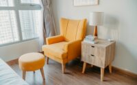 yellow armchair and stool beside wooden nightstand by the wall near glass window and bed